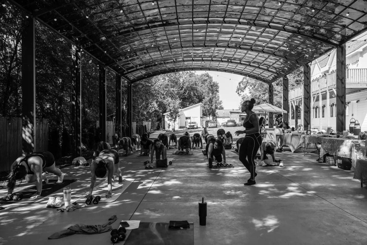 A group of people practice yoga under a large, open-sided, covered pavilion. An instructor stands at the front, observing participants who are in a tabletop yoga pose. The scene is in black and white. Huntersville event venue