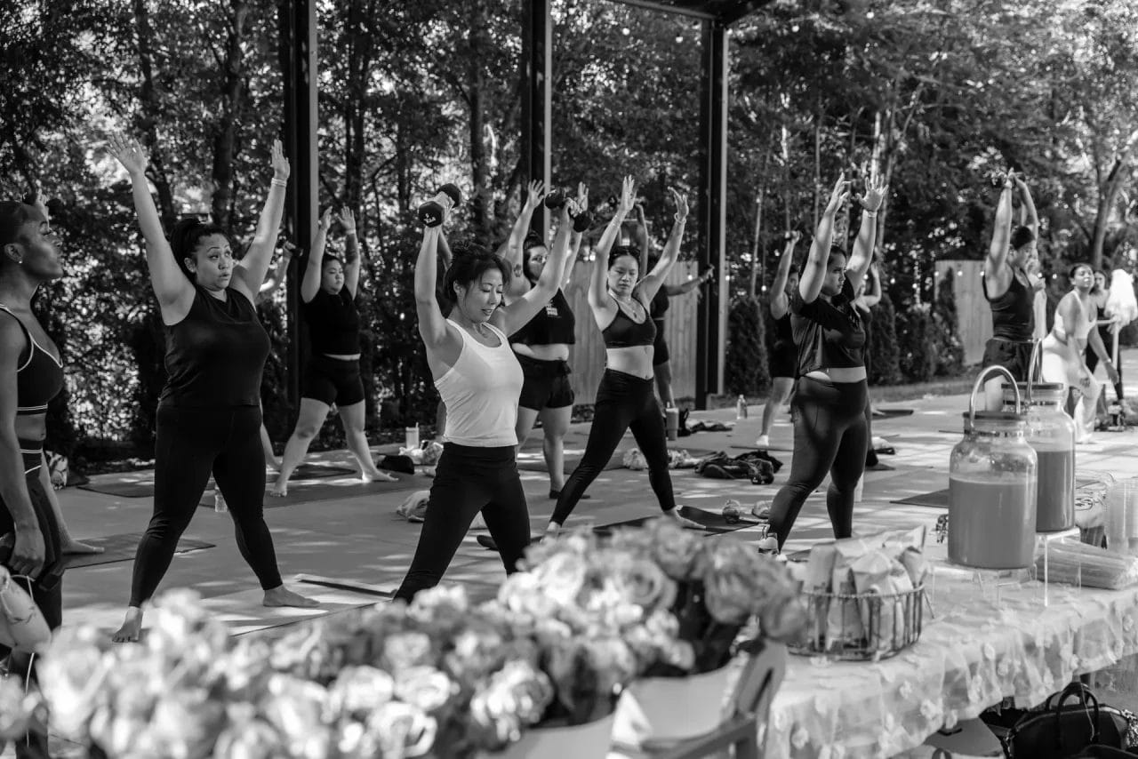 A group of people in athletic wear perform a yoga or fitness class outdoors, raising their arms overhead. In the foreground, a table with flowers and drink dispensers is visible. The setting is surrounded by trees. Huntersville event venue