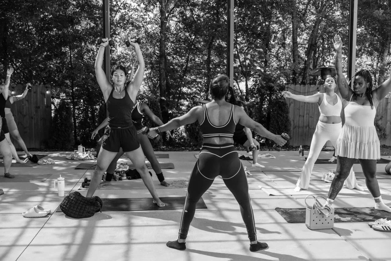 A group of women participate in an outdoor fitness class, exercising on yoga mats under a covered space with sunlight filtering through trees in the background. Huntersville event venue