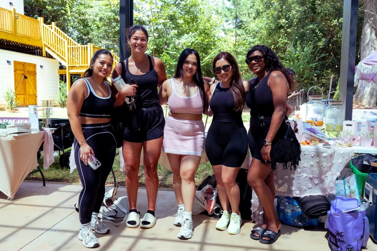 Five women in athletic wear stand smiling together outdoors by a decorated table, with trees and a yellow-railed building in the background. They appear to be at a casual gathering or party. Huntersville event venue