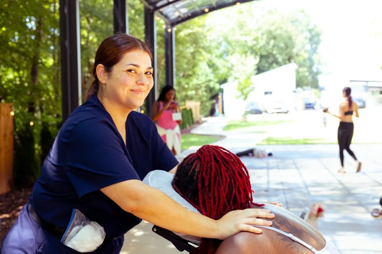 A smiling massage therapist in navy scrubs gives a seated outdoor massage to a client with red braids. Other people and greenery are visible in the sunlit background. Huntersville event venue
