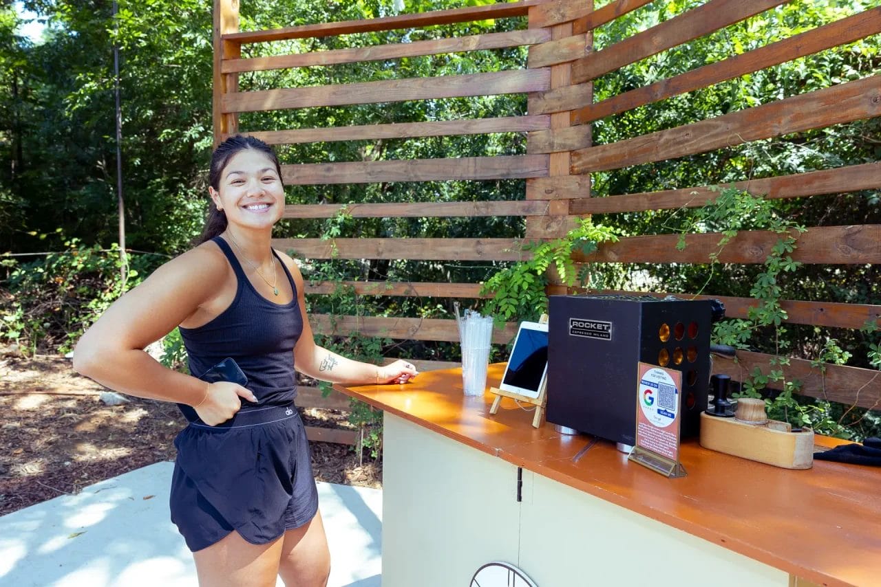 A smiling woman in athletic wear stands at an outdoor refreshment stand with a wooden slatted backdrop, surrounded by greenery and sunlight. Cups, a tablet, and a payment terminal are visible on the counter. Huntersville event venue