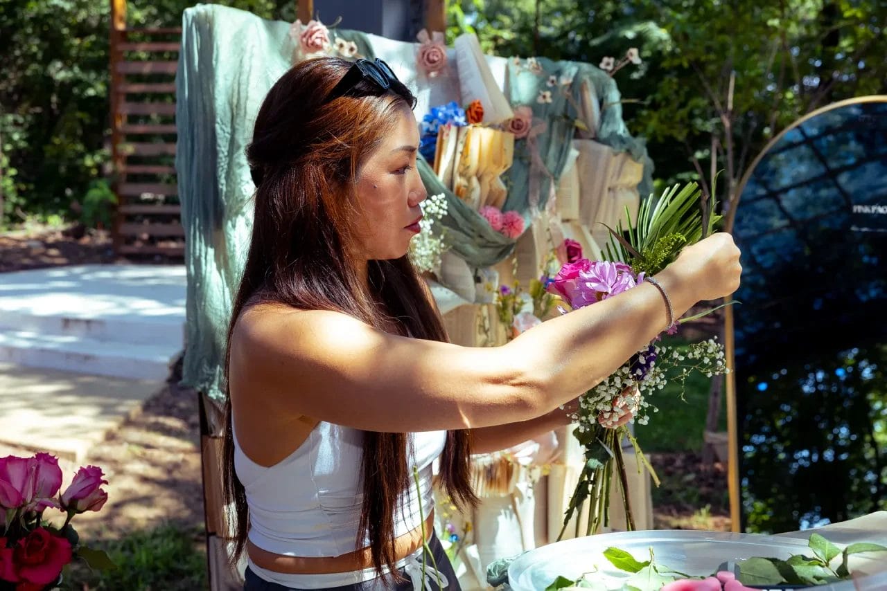 A woman with long brown hair arranges a bouquet of flowers outdoors, surrounded by various floral decorations and greenery. She is focused on her work, wearing a white tank top and sunglasses on her head. Huntersville event venue