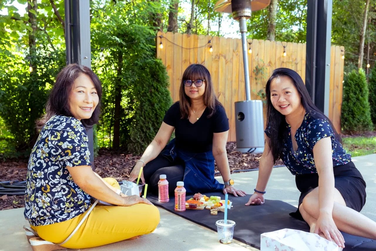 Three women sit on a mat and pavement outdoors, enjoying food and drinks together. They are smiling at the camera, surrounded by greenery, with a wooden fence and patio heater in the background. Huntersville event venue