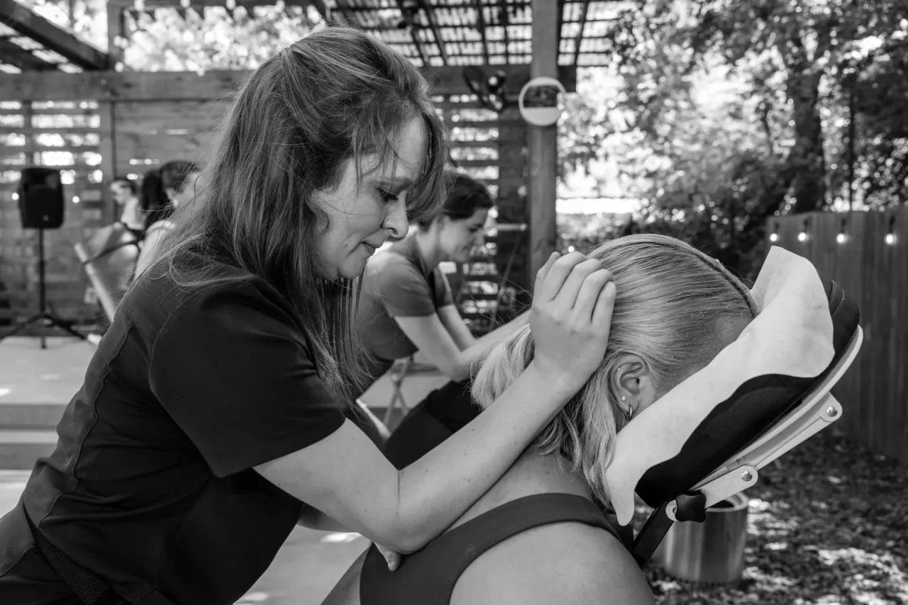 A woman gives a seated head and neck massage to another woman outdoors, while other people receive similar massages in the background. The scene is shaded by a wooden pergola. Huntersville event venue