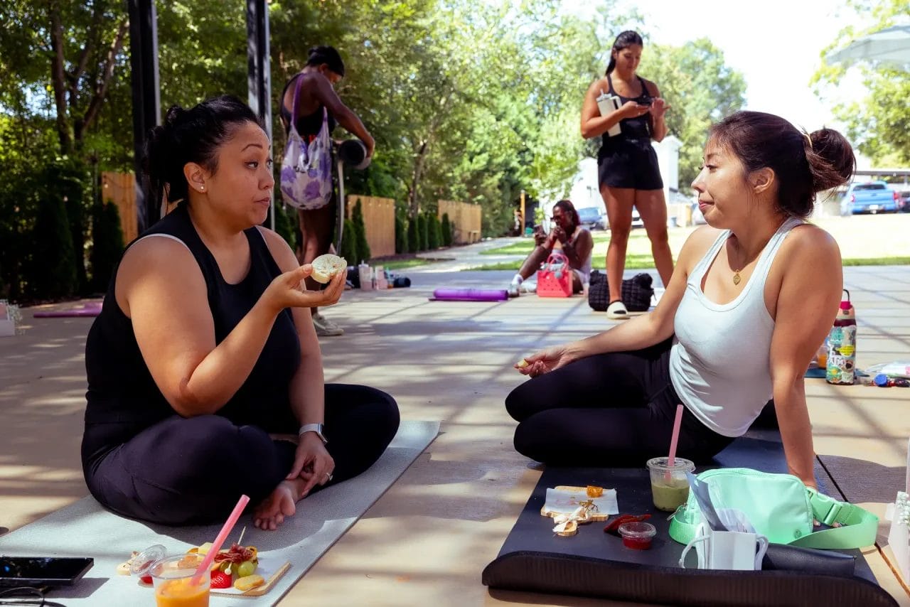 Two women sit on yoga mats outdoors, enjoying drinks and snacks while talking. Other people with yoga mats and water bottles are in the background under trees, suggesting a relaxed group setting. Huntersville event venue