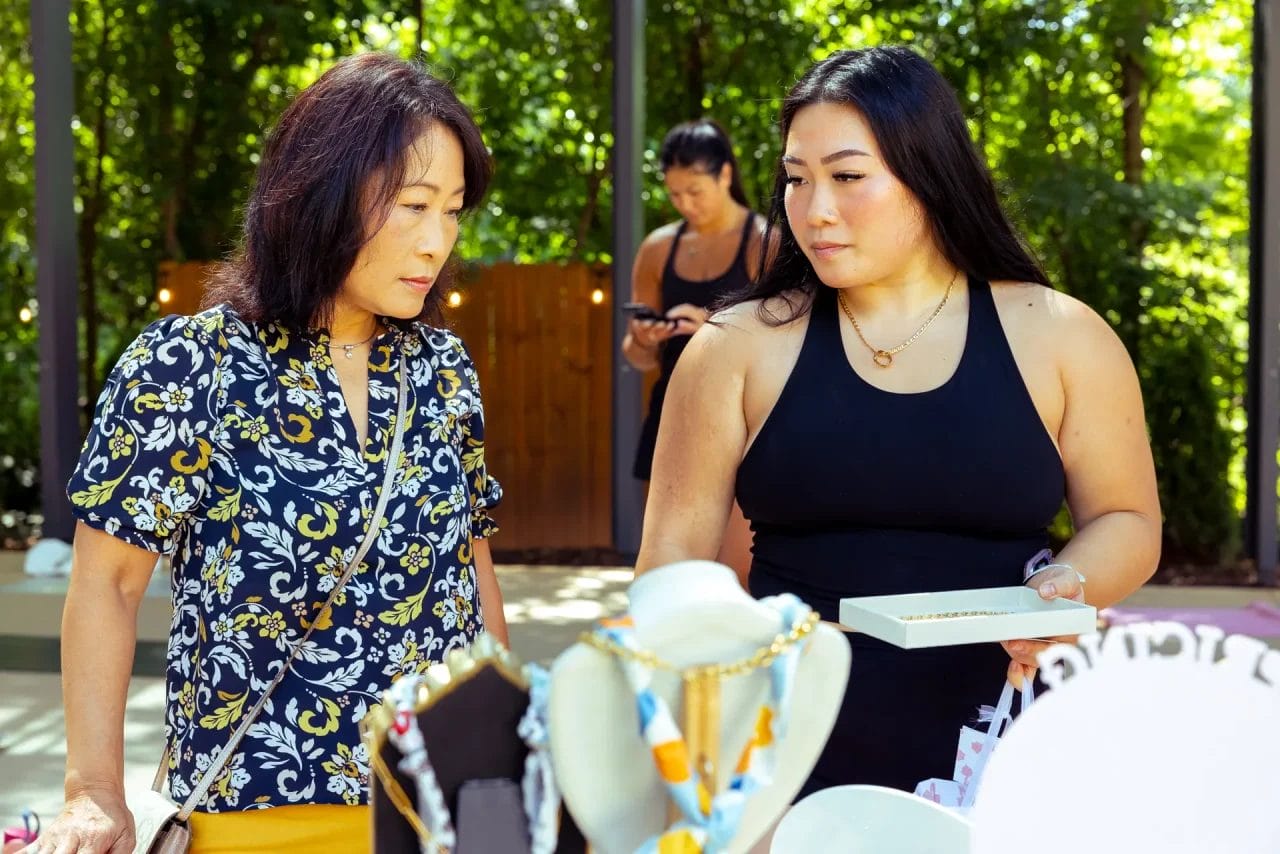 Two women stand at an outdoor jewelry stall, thoughtfully looking at displayed necklaces and accessories. One woman holds a white box while the other observes. A third person is blurred in the background among trees. Huntersville event venue