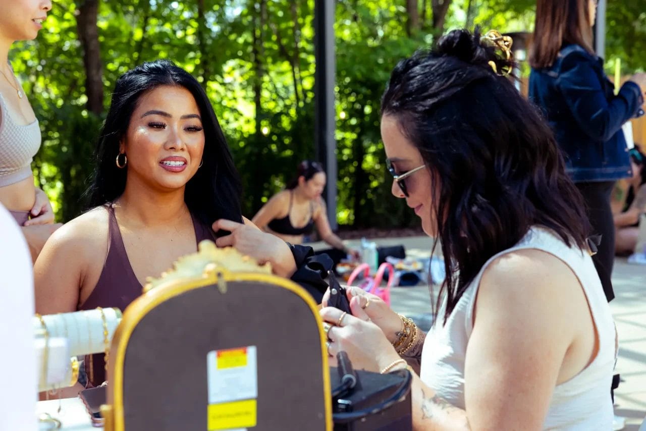 Two women sit outdoors at a table with accessories. One smiles and touches her hair, while the other works with her hands. People are blurred in the background, and trees provide a green backdrop. Huntersville event venue