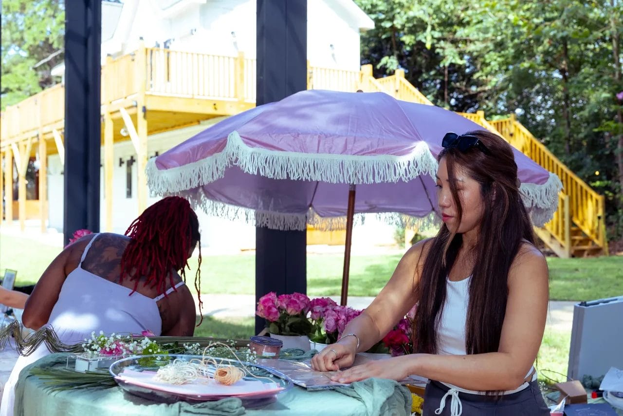 Two women work at an outdoor table covered with craft supplies and flowers. A pink fringed umbrella provides shade, and a yellow wooden building with stairs is visible in the background. Huntersville event venue