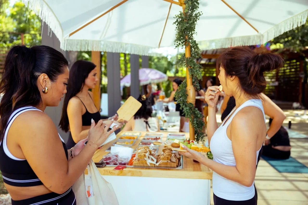 Women in activewear gather around an outdoor food stand under a white umbrella, sampling snacks and chatting in a sunny, garden-like setting. One woman holds a plate of food while another writes on a notepad. Huntersville event venue