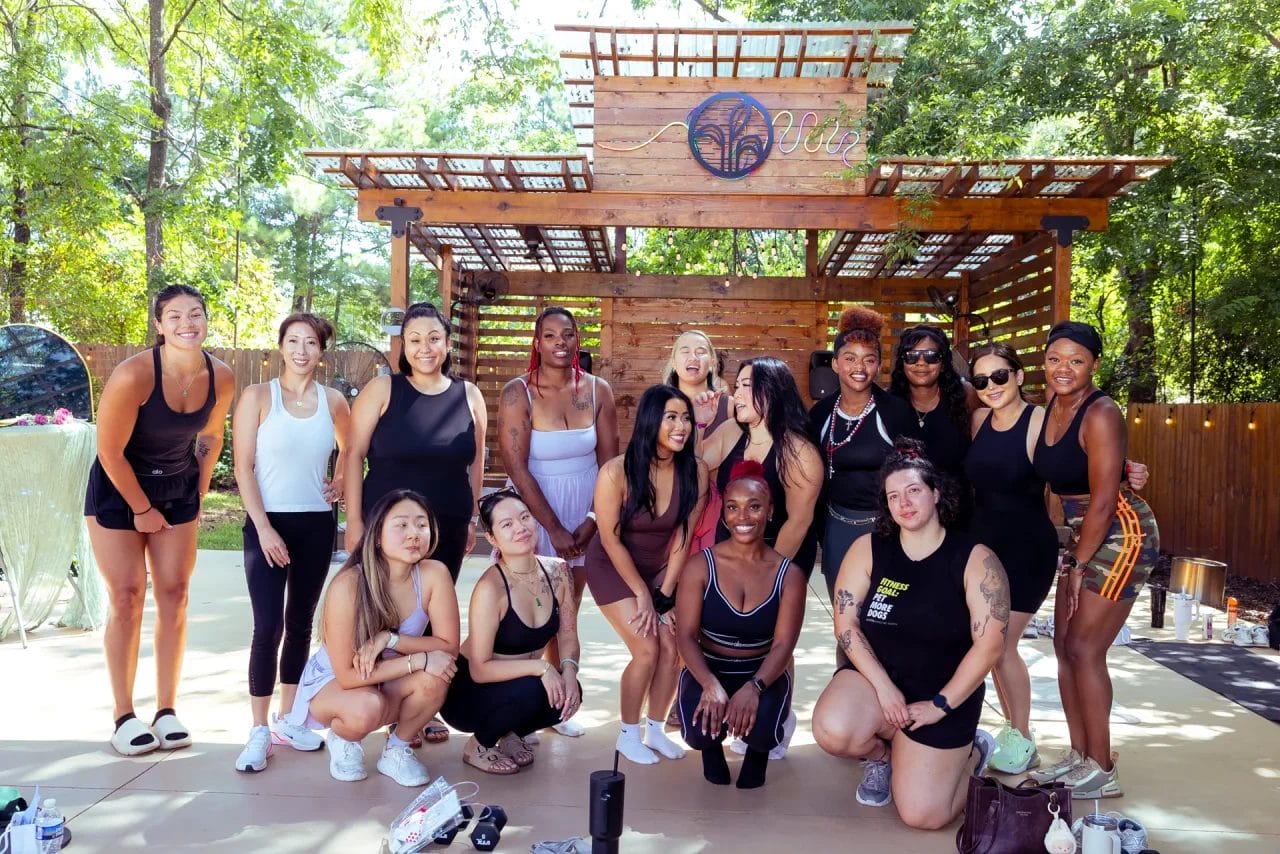 A group of women in athletic wear pose and smile together outdoors on a sunny day, standing and kneeling in front of a wooden pergola surrounded by trees. Huntersville event venue