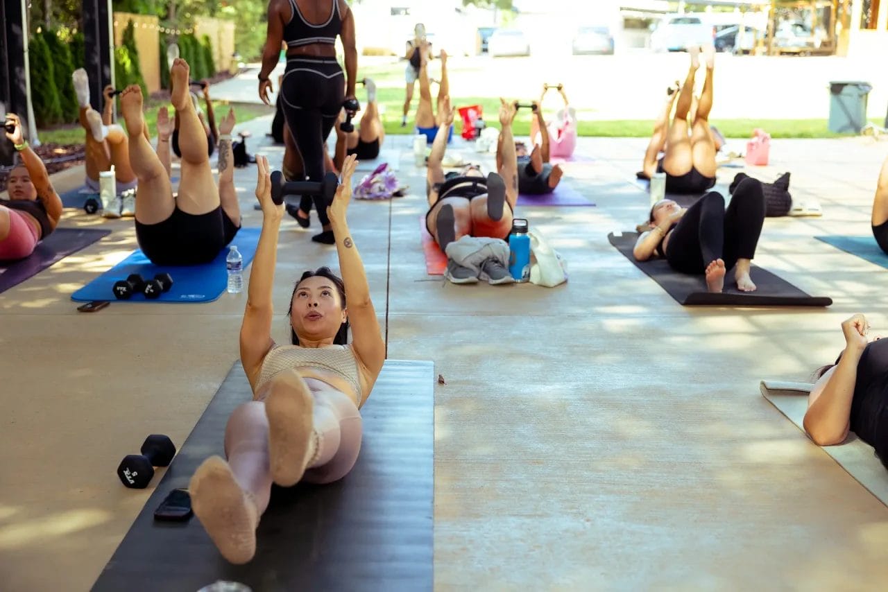 A group of people exercise on yoga mats outdoors, lying on their backs with arms and legs raised, following an instructor at the front. Sunlight shines on the scene, and water bottles are scattered nearby. Huntersville event venue