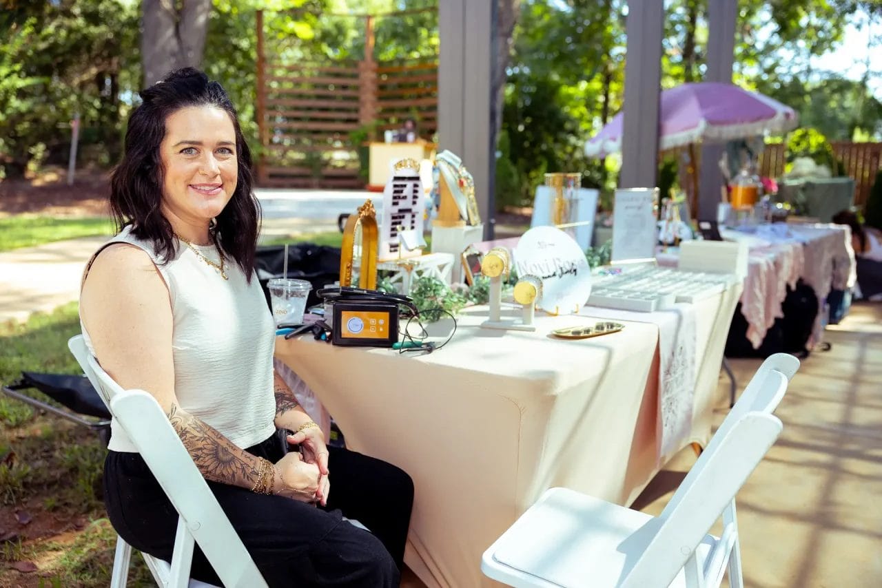 A woman with dark hair and tattoos sits smiling at a table displaying jewelry and other items at an outdoor market under a canopy, with trees and sunlight in the background. Huntersville event venue