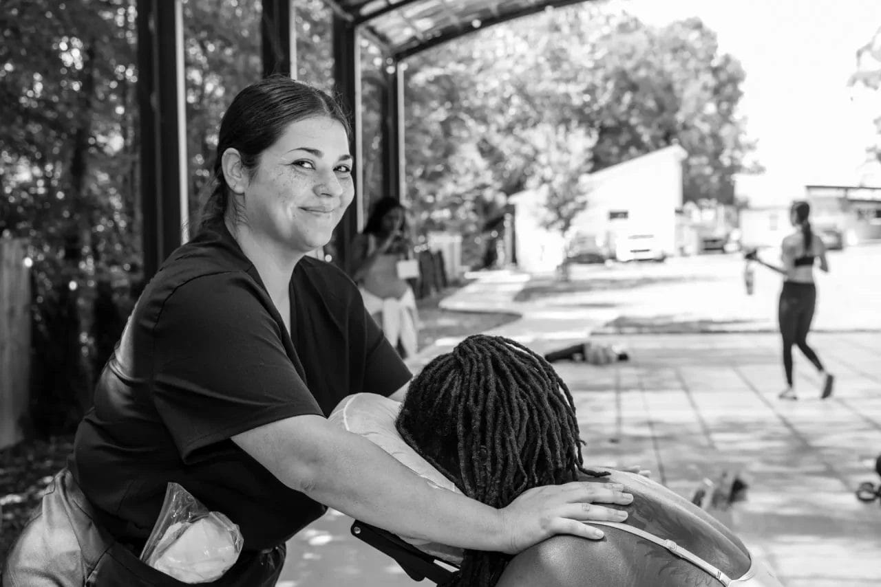 A woman smiles at the camera while giving a seated massage to another person outdoors under a covered area, with other people visible in the background. Huntersville event venue