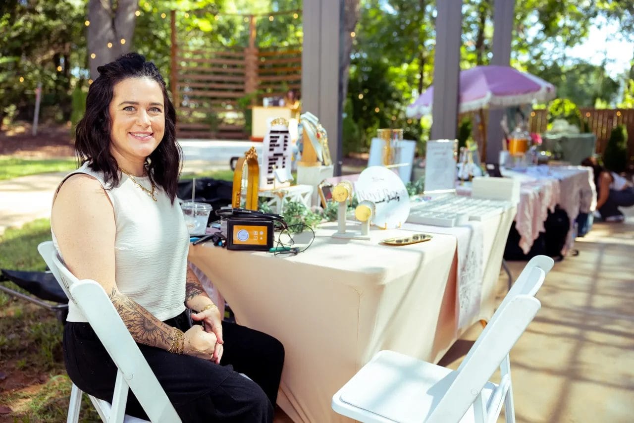 A woman with dark hair and tattoos sits smiling at a table with art supplies and decorations at an outdoor event, with other decorated tables and greenery visible in the background. Huntersville event venue