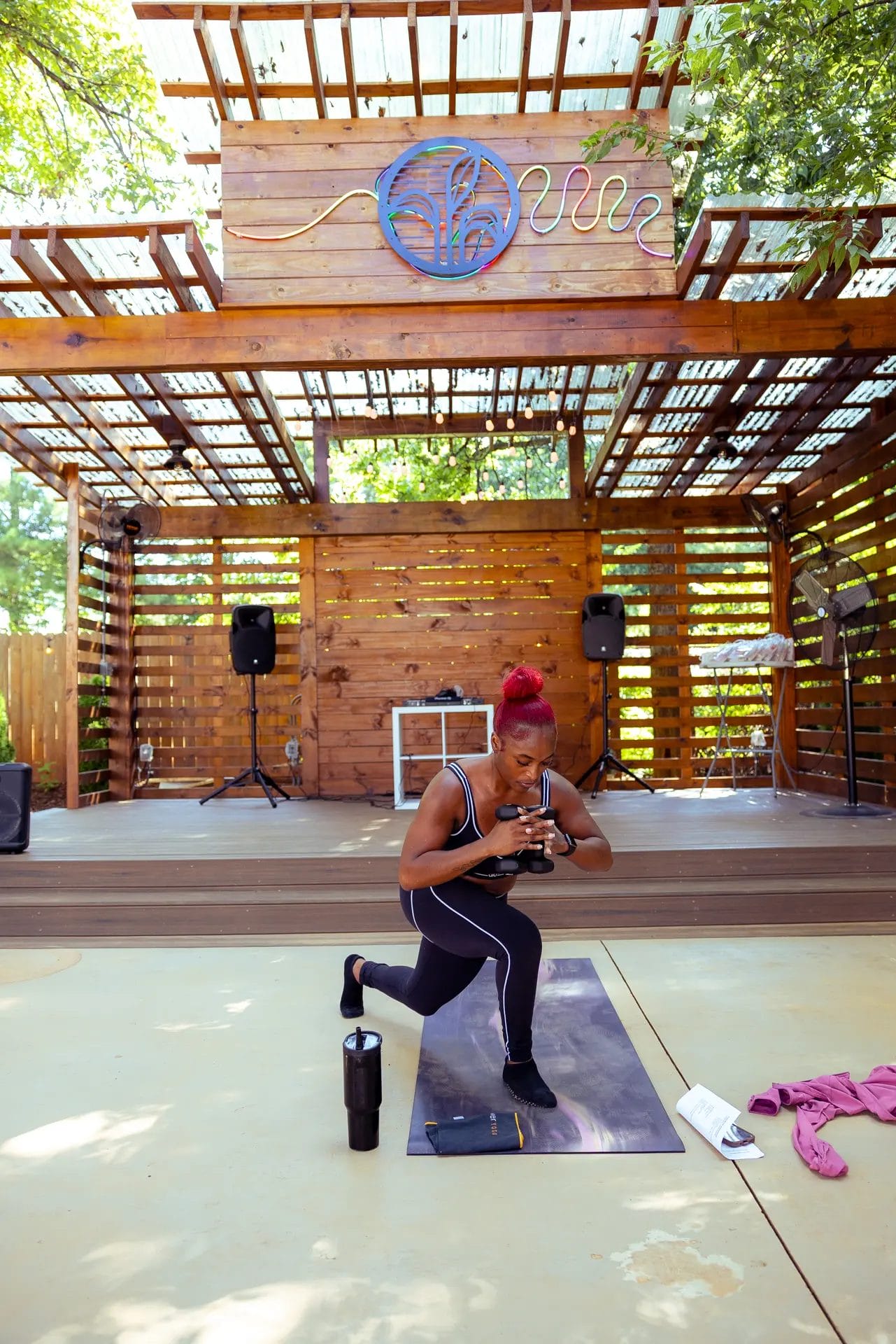 A woman with red hair exercises on a yoga mat outdoors under a wooden pavilion, performing a lunge while looking at her phone. Fitness gear, a towel, water bottle, and speakers are visible around her. Huntersville event venue