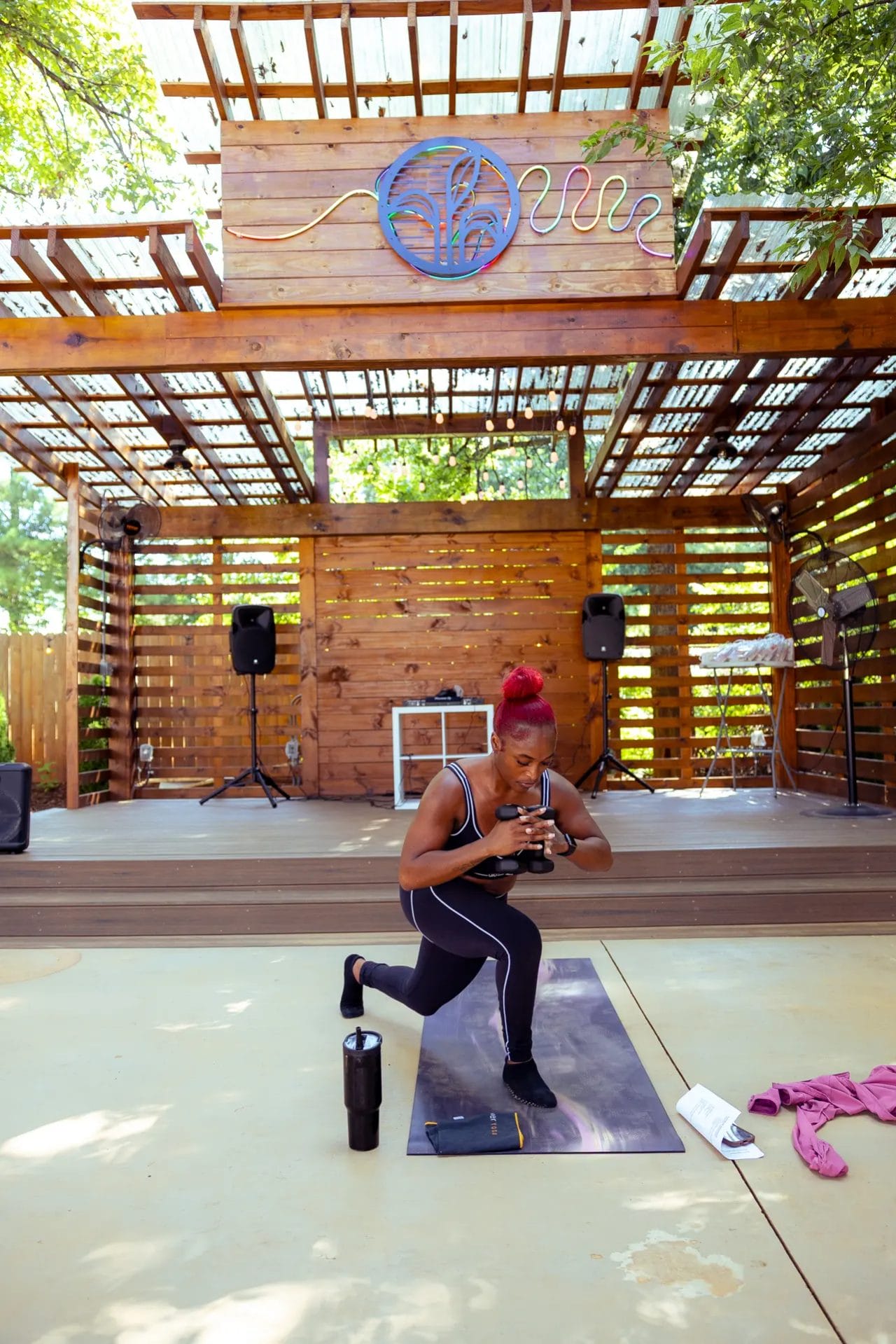 Rent a Yoga Space near Charlotte, NC — Covered Studio & Outdoor Options 9 A woman with red hair exercises on a yoga mat outdoors under a wooden pavilion, performing a lunge while looking at her phone. Fitness gear, a towel, water bottle, and speakers are visible around her. Huntersville event venue