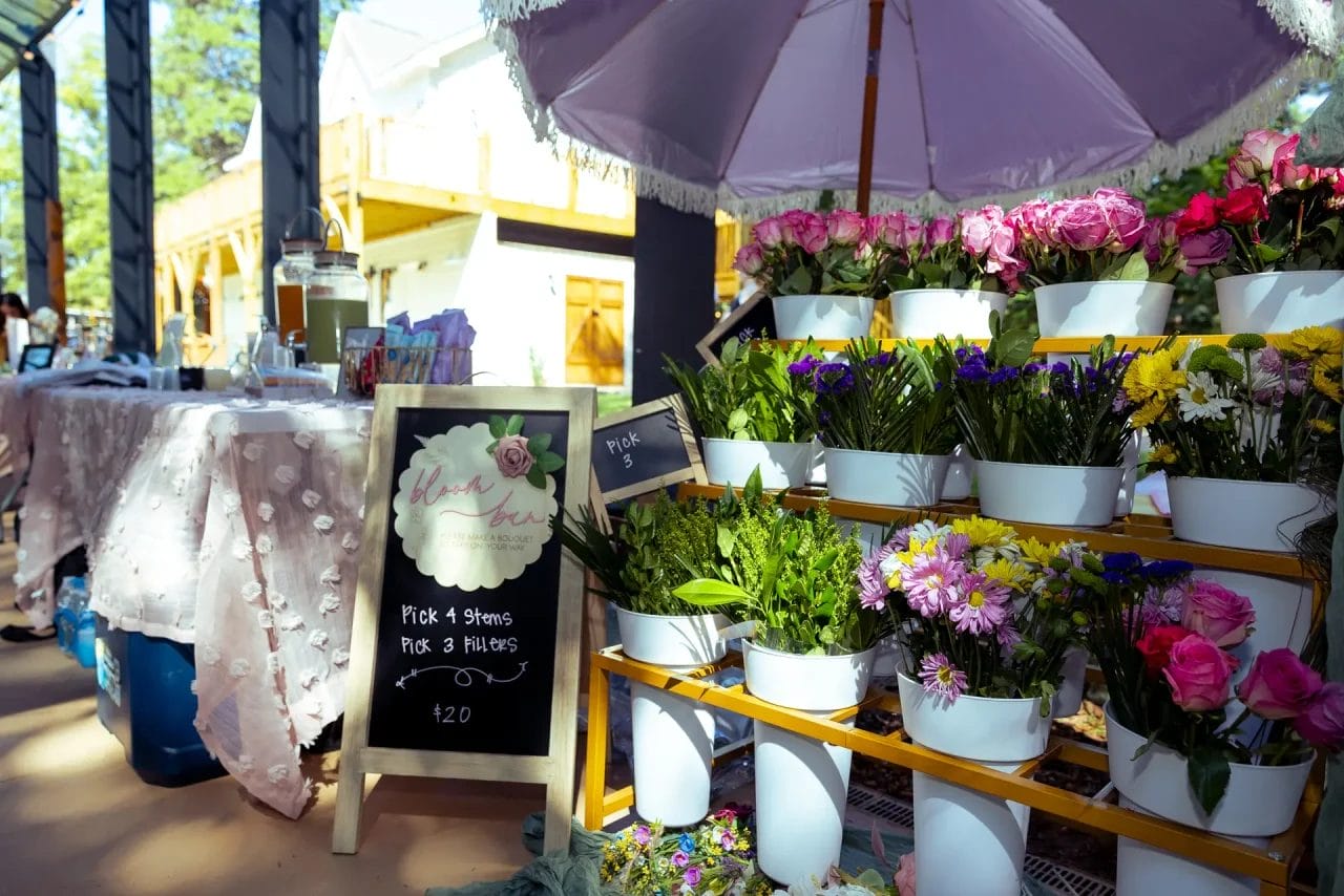 A flower stand at an outdoor market features rows of colorful flowers in white pots, a chalkboard sign with prices, and a lavender umbrella providing shade on a sunny day. Huntersville event venue