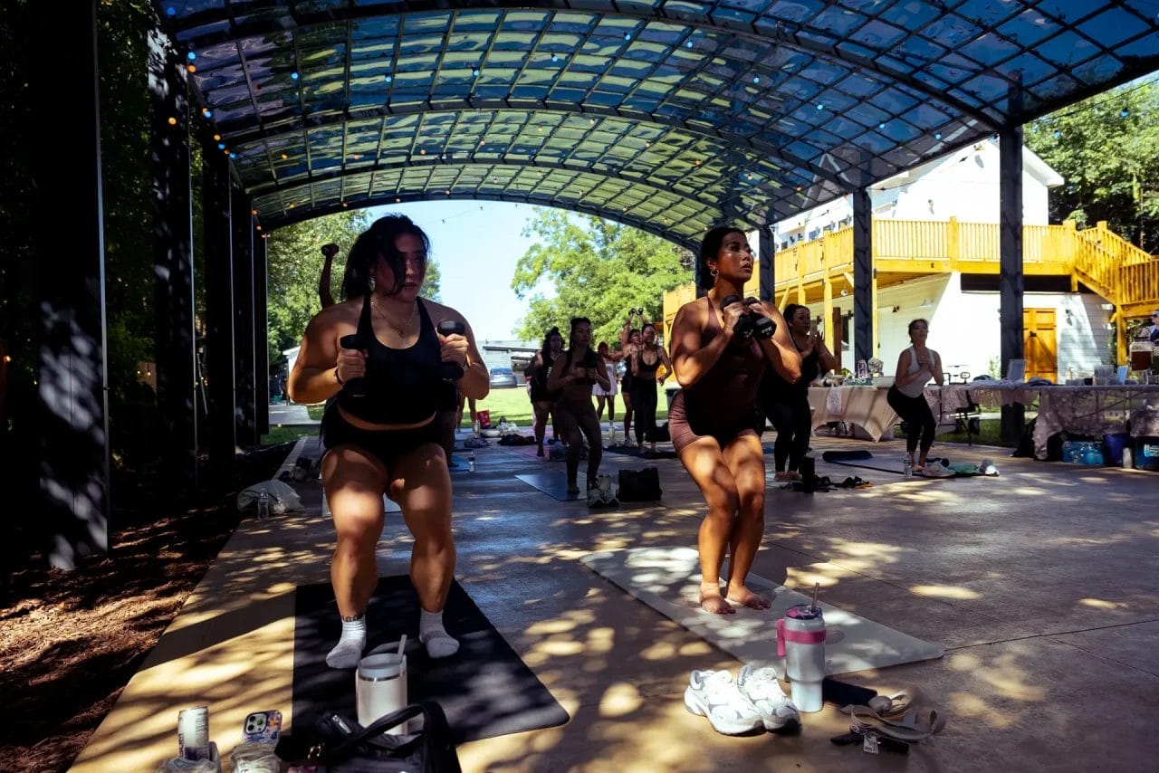 A group of people in workout clothes exercise together on yoga mats under a large, open-roofed pavilion with sunlight streaming through, surrounded by trees and nearby buildings. Huntersville event venue