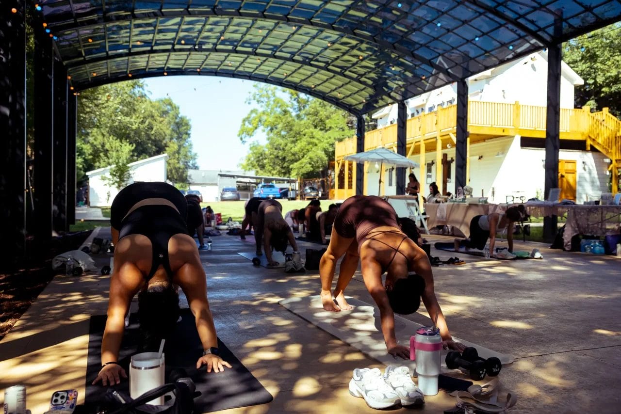 People are practicing yoga on mats under a large open-air pavilion. Most are in a downward dog pose. Sunlight filters through the roof. Shoes, water bottles, and towels are beside them. A building is visible in the background. Huntersville event venue