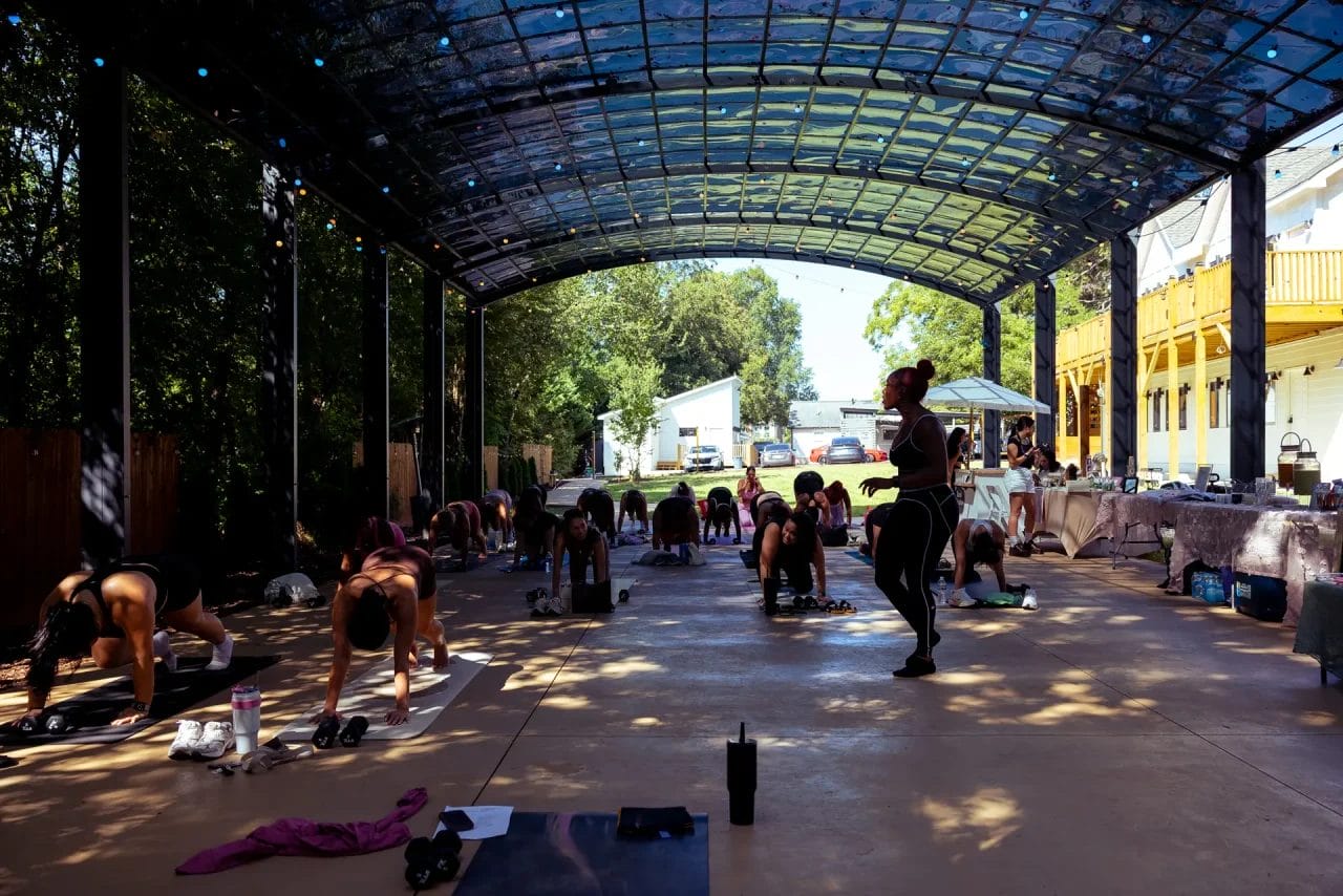 A group of people practice yoga on mats under a large, open-air canopy, with an instructor standing and guiding them; sunlight filters in, and trees and buildings are visible outside. Huntersville event venue