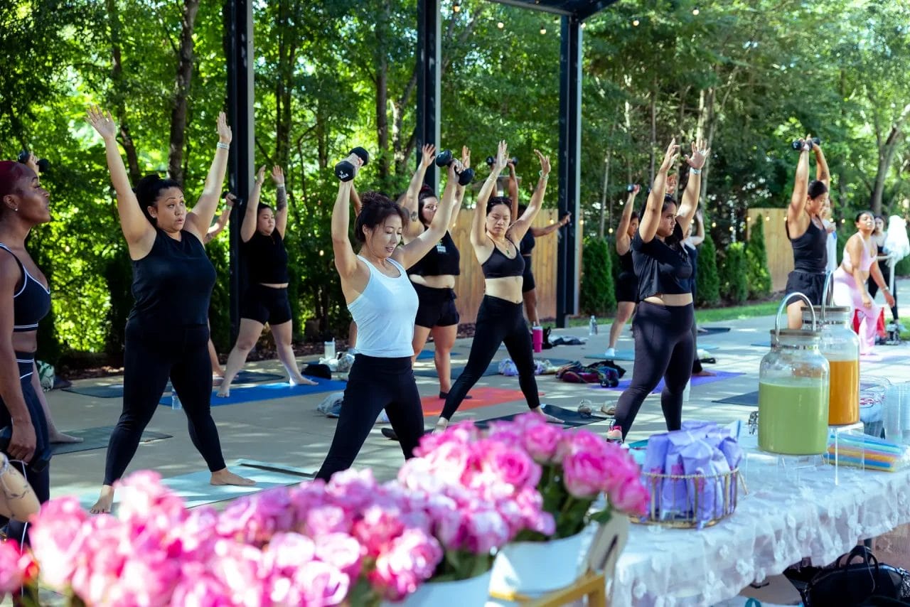 A group of women in workout clothes exercise with dumbbells outdoors on yoga mats, surrounded by trees. In the foreground, a table displays pink flowers, drinks, and small gift bags. Huntersville event venue