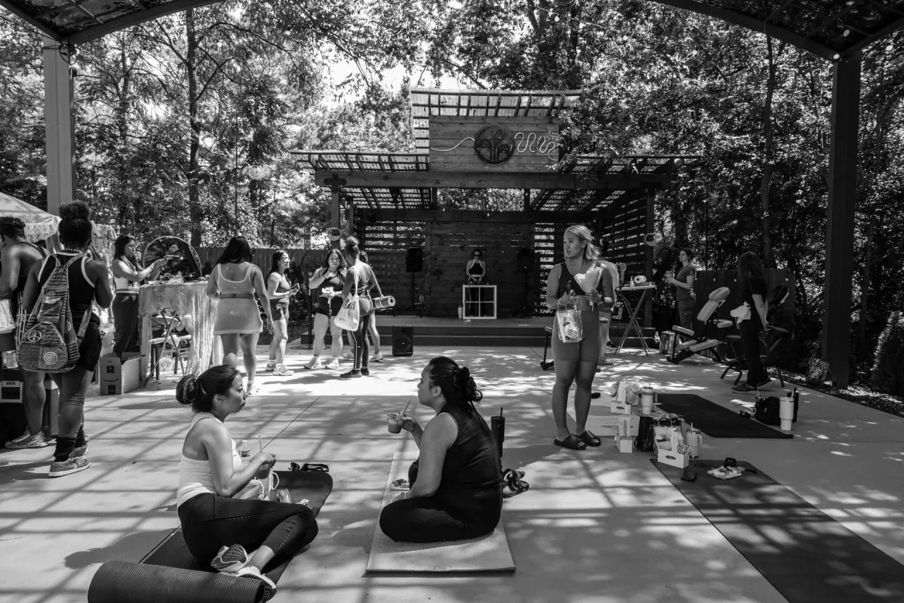 A group of people gathers in an outdoor, covered pavilion. Some sit on yoga mats talking, while others stand or walk around. A DJ plays music on a stage in the background, surrounded by trees and dappled sunlight. Huntersville event venue