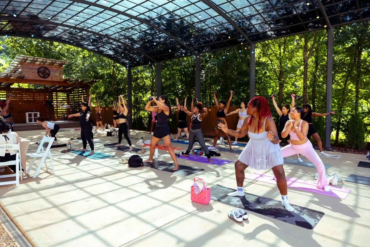A diverse group of people practice yoga on mats under a large, glass-covered outdoor pavilion surrounded by trees. Most participants are in active poses, with sunlight filtering through the canopy. Huntersville event venue