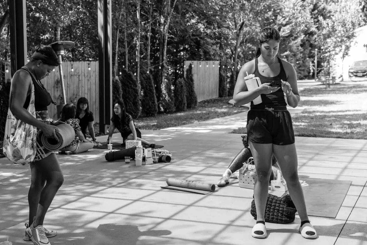 A group of people are outdoors under a shaded structure. One person stands checking her phone, holding a water bottle, while others sit on yoga mats and talk. Sunlight filters through trees and a fence lines the background. Huntersville event venue