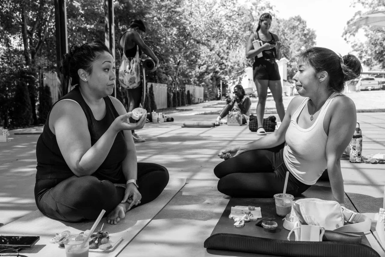 Two women in workout clothes sit cross-legged on mats outdoors, chatting and eating snacks. Other people stand and sit in the background. Food and drinks are placed on the mats. The scene is relaxed and casual, with trees nearby. Huntersville event venue