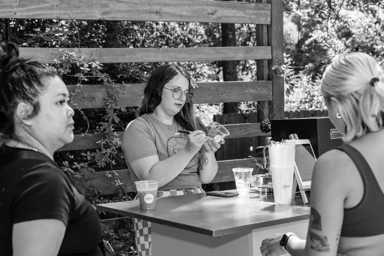 Three women interact at an outdoor booth; one stands behind the counter taking notes, while the other two stand in front. Cups and straws are on the counter. The setting is shaded by trees and a wooden fence. Huntersville event venue