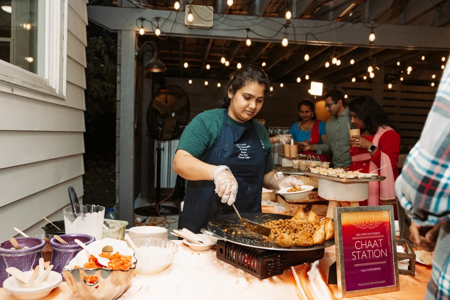 A person is preparing food at an outdoor chaat station under string lights, transforming the first birthday party venue into a vibrant culinary scene. Various ingredients and dishes are displayed on the table, as guests gather around, captivated by the delightful preparation. Huntersville event venue