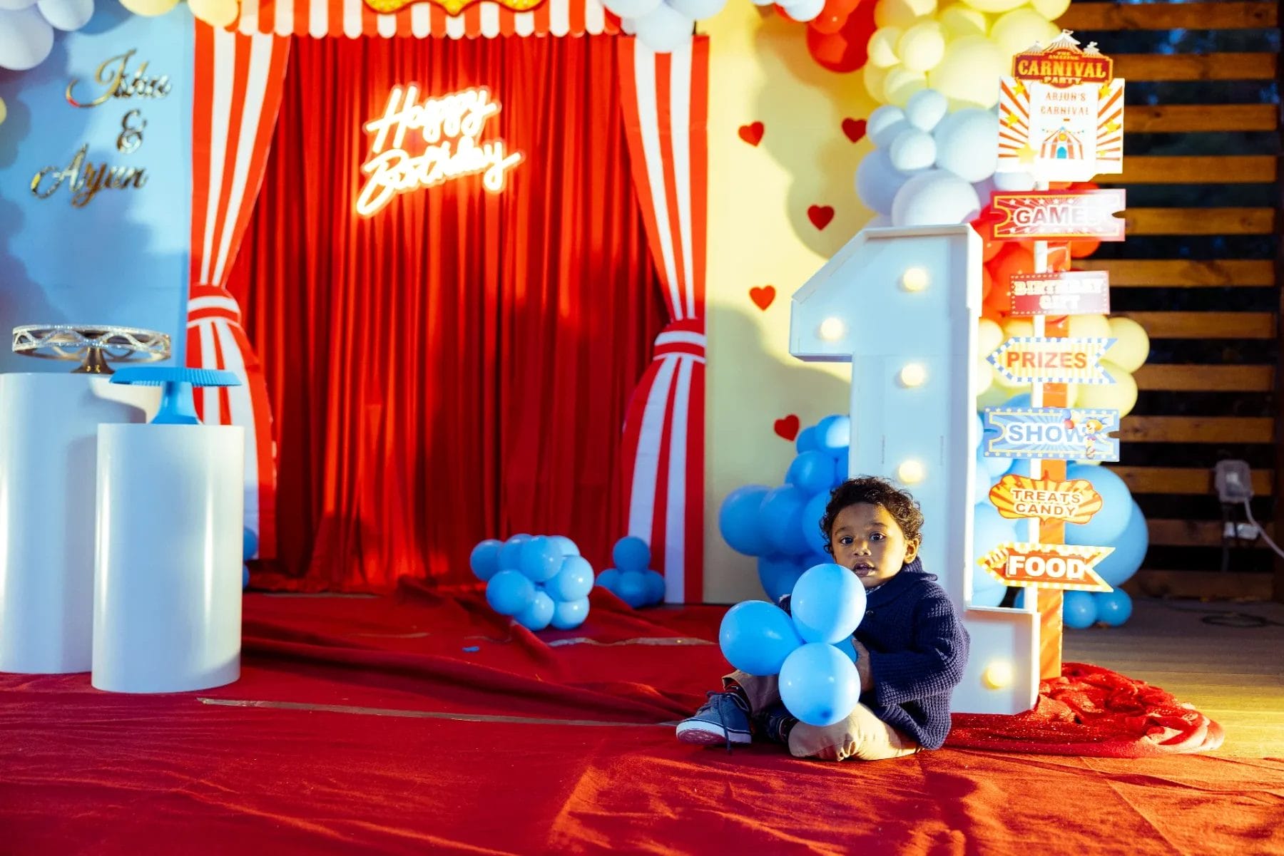 A toddler sits on a red carpet holding a blue balloon, surrounded by colorful carnival-themed decorations perfect for a first birthday party venue. A large number one stands proudly amid balloons, while a red curtain backdrop showcases "Happy Birthday" in vibrant neon lights. Huntersville event venue