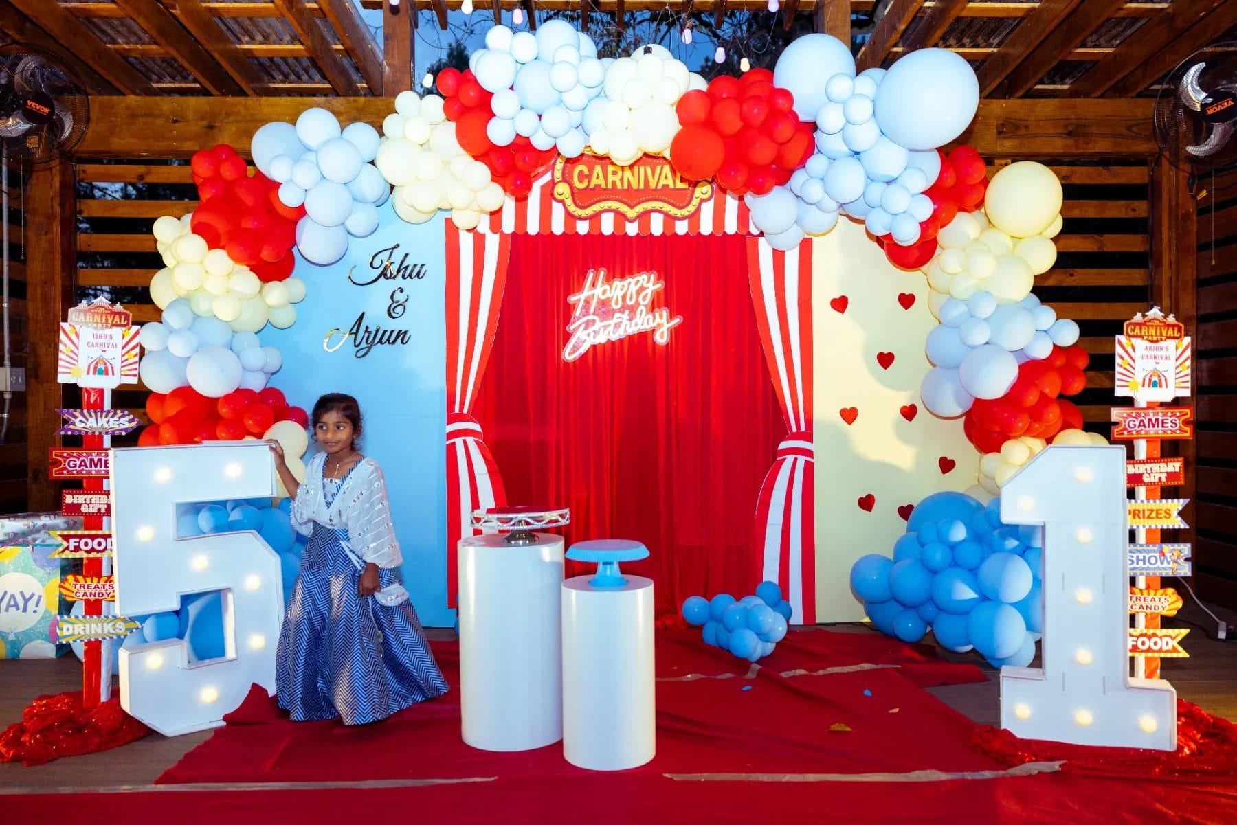 A festive first birthday party venue with a carnival theme features clusters of white, red, and blue balloons. A young girl stands next to large illuminated numbers "5" and "1" on a red carpet. A "Happy Birthday" sign hangs above a vibrant red curtain backdrop, setting the perfect celebratory scene. Huntersville event venue