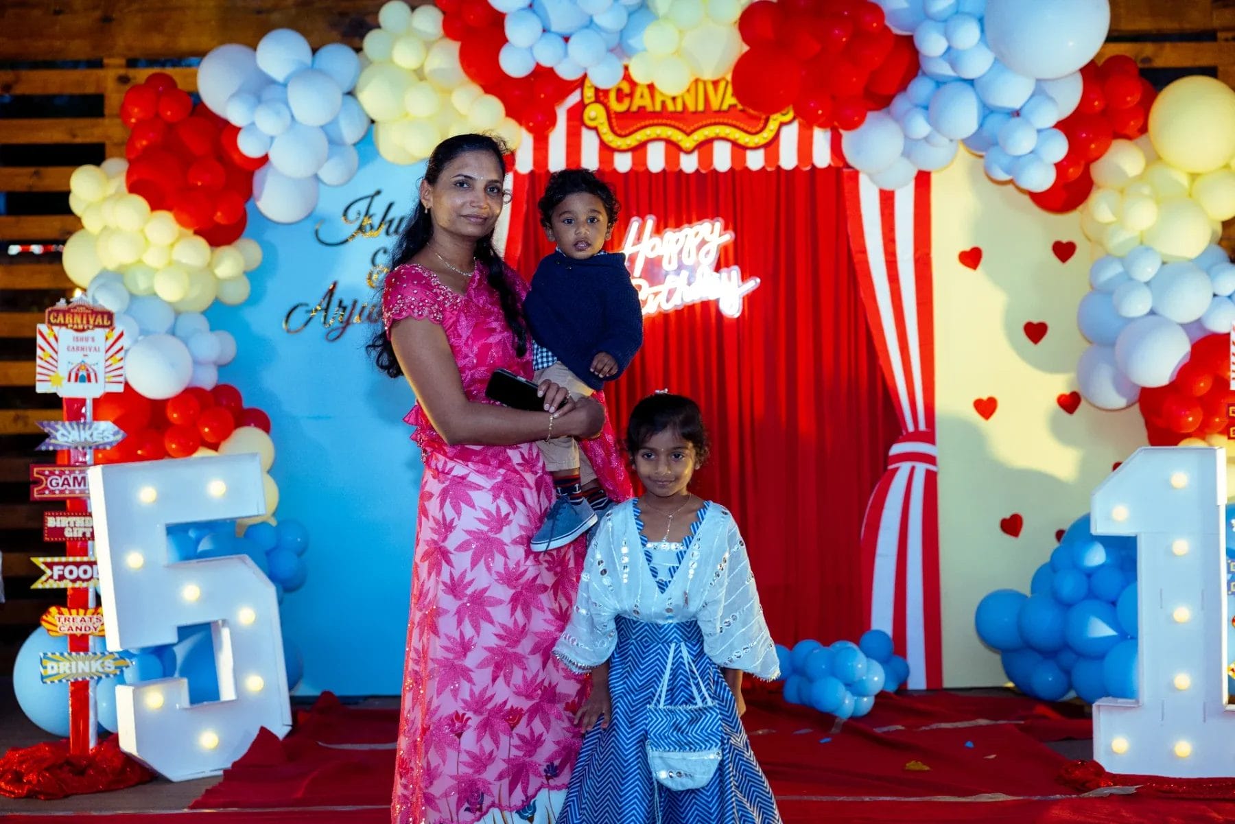 A woman in a pink dress holds a young boy beside a girl in a blue dress at a festive first birthday party venue. The carnival-themed celebration features colorful balloons and a red curtain backdrop displaying "Happy Birthday. Huntersville event venue