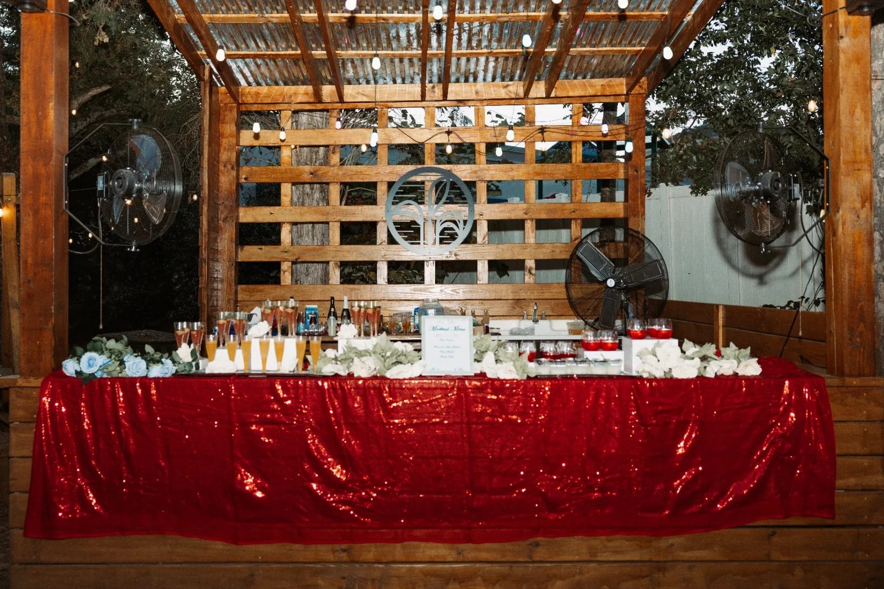 A rustic outdoor bar setup serves as the perfect first birthday party venue, with a wooden frame and string lights overhead. A red sequined tablecloth adorns the bar, showcasing an array of drink glasses and bottles. Decorative silver fans are mounted on the sides, framed by trees in the background. Huntersville event venue