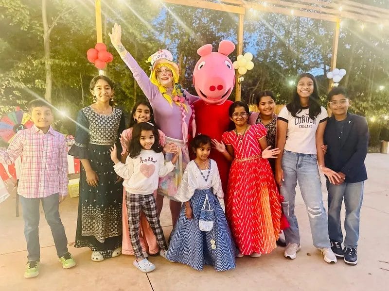 A group of children and two adults in colorful costumes pose happily together outdoors. One adult is dressed as a clown, and the other is in a red costume with a large pink head. String lights and balloons are in the background. Huntersville event venue