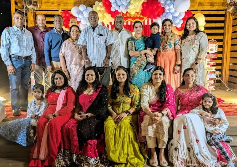 A family group photo with several adults and children posing in front of a festive backdrop decorated with red and white balloons and the word "Carnival." The group is dressed in colorful attire, with smiles and a warm, celebratory atmosphere. Huntersville event venue