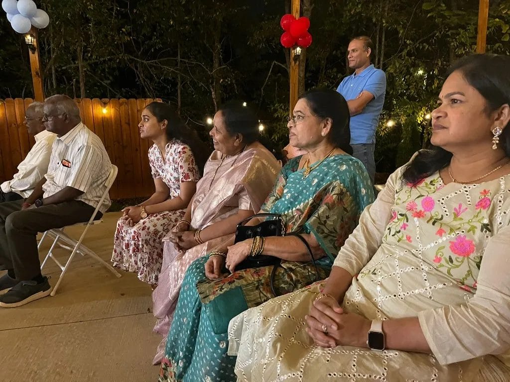 A group of people sitting in a row, some wearing traditional clothing. Two women and a girl are in floral or colorful dresses, while two men and another woman wear more formal attire. Decorative red and blue balloons are above them, with trees in the background. Huntersville event venue