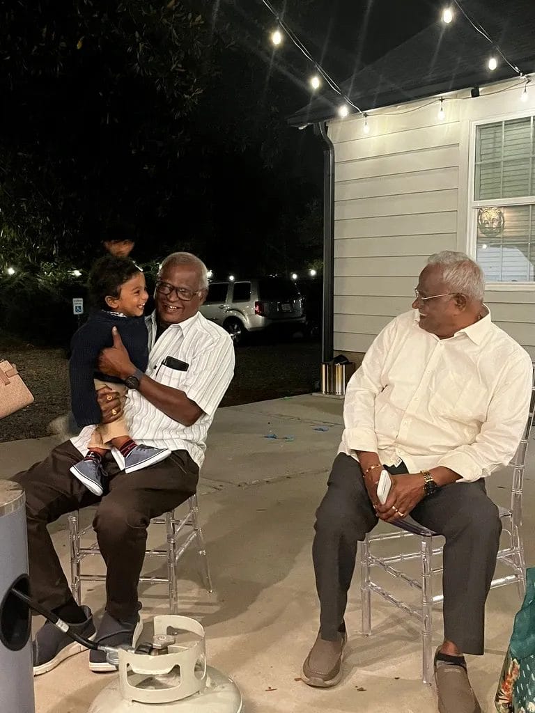 Two elderly men sit on chairs outside. One man, wearing a striped shirt, holds a young child who is smiling. The other man, in a white shirt, watches them. String lights illuminate the scene, and a house is visible in the background. Huntersville event venue