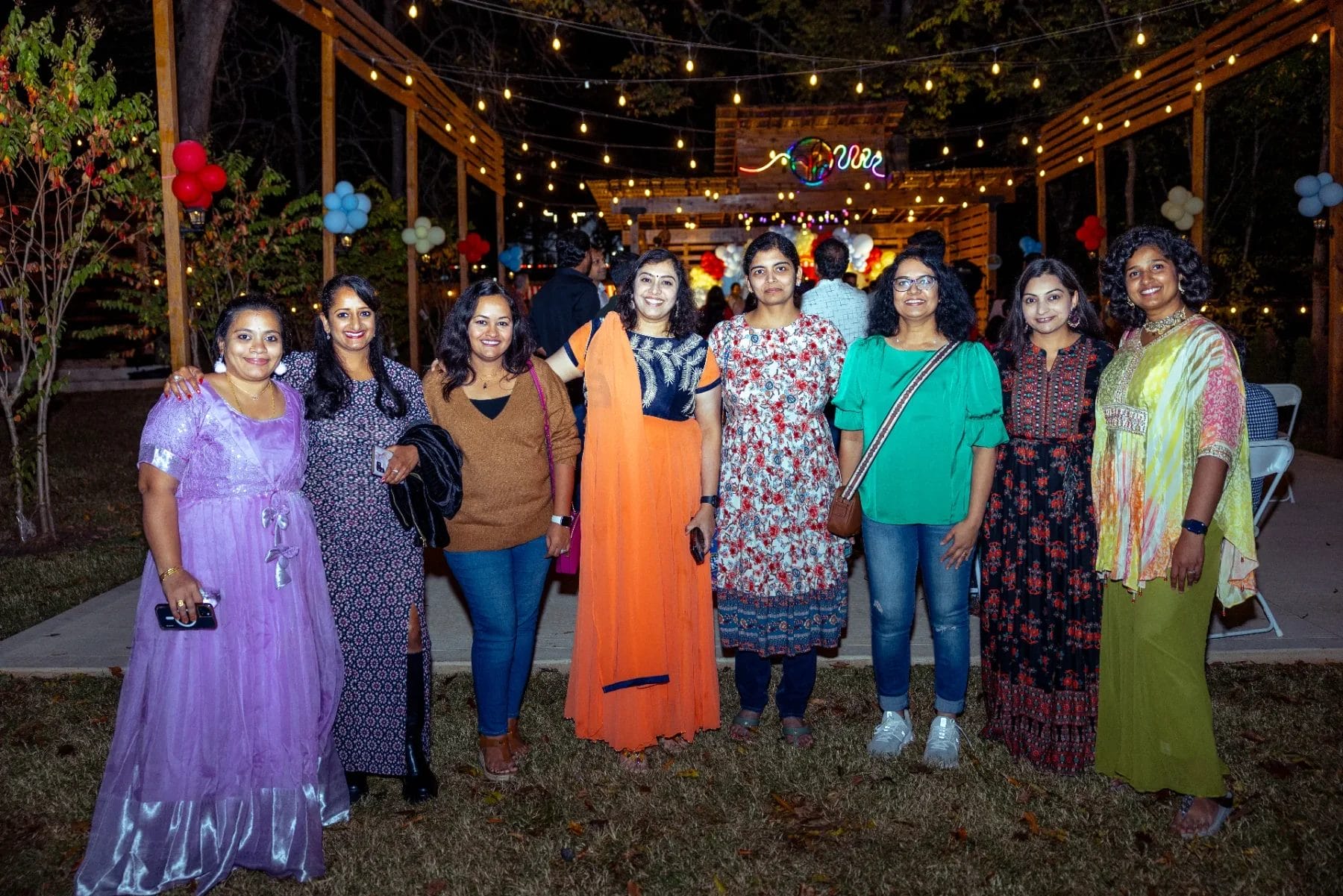 A group of eight people stands outdoors at night, smiling for a photo at a festive first birthday party venue. They're dressed in colorful outfits, with festive lights and balloons in the background, while trees and a wooden structure complete the scene. Huntersville event venue