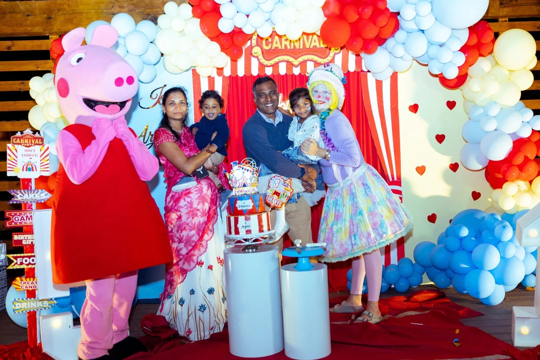 A family of four poses with a person in a pink costume and a clown at the perfect first birthday party venue. They stand in front of a vibrant carnival-themed backdrop adorned with balloons and decorations, while a table with an eye-catching cake and treats takes center stage. Huntersville event venue