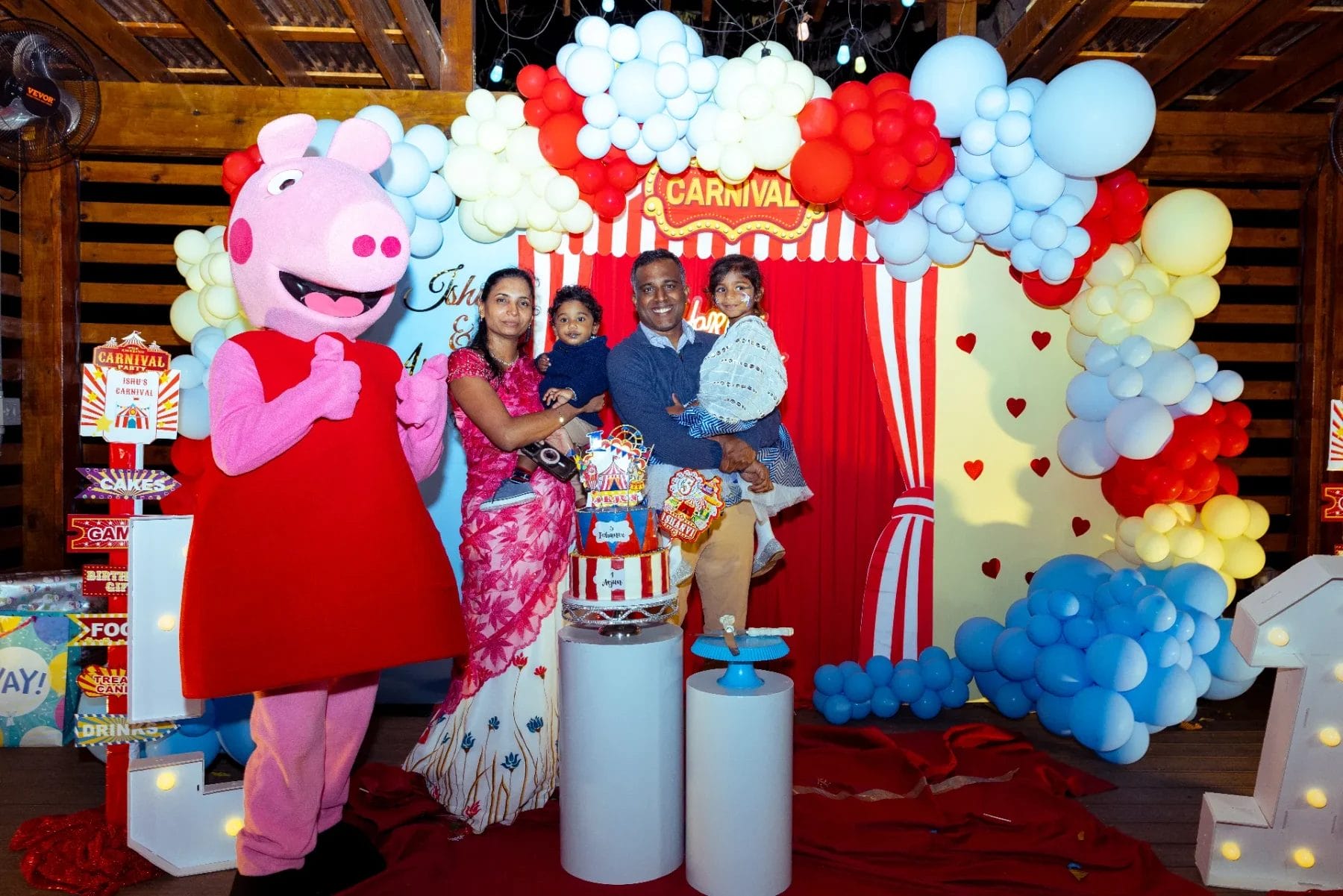 A family of four poses at a vibrant first birthday party venue. They stand beside a large cartoon character, surrounded by a colorful balloon backdrop of red, white, and blue. In the foreground, a decorated birthday cake sits on the table, completing the festive carnival atmosphere. Huntersville event venue