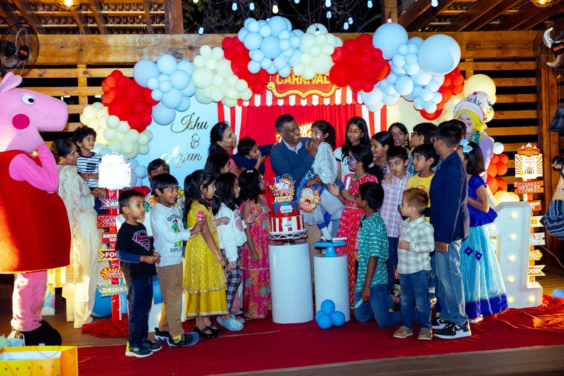 Children gather around a man holding a baby at a colorful first birthday party venue, adorned with balloons and cartoon character decorations. Bright lights illuminate the scene, and a large cake stands proudly before them. Huntersville event venue