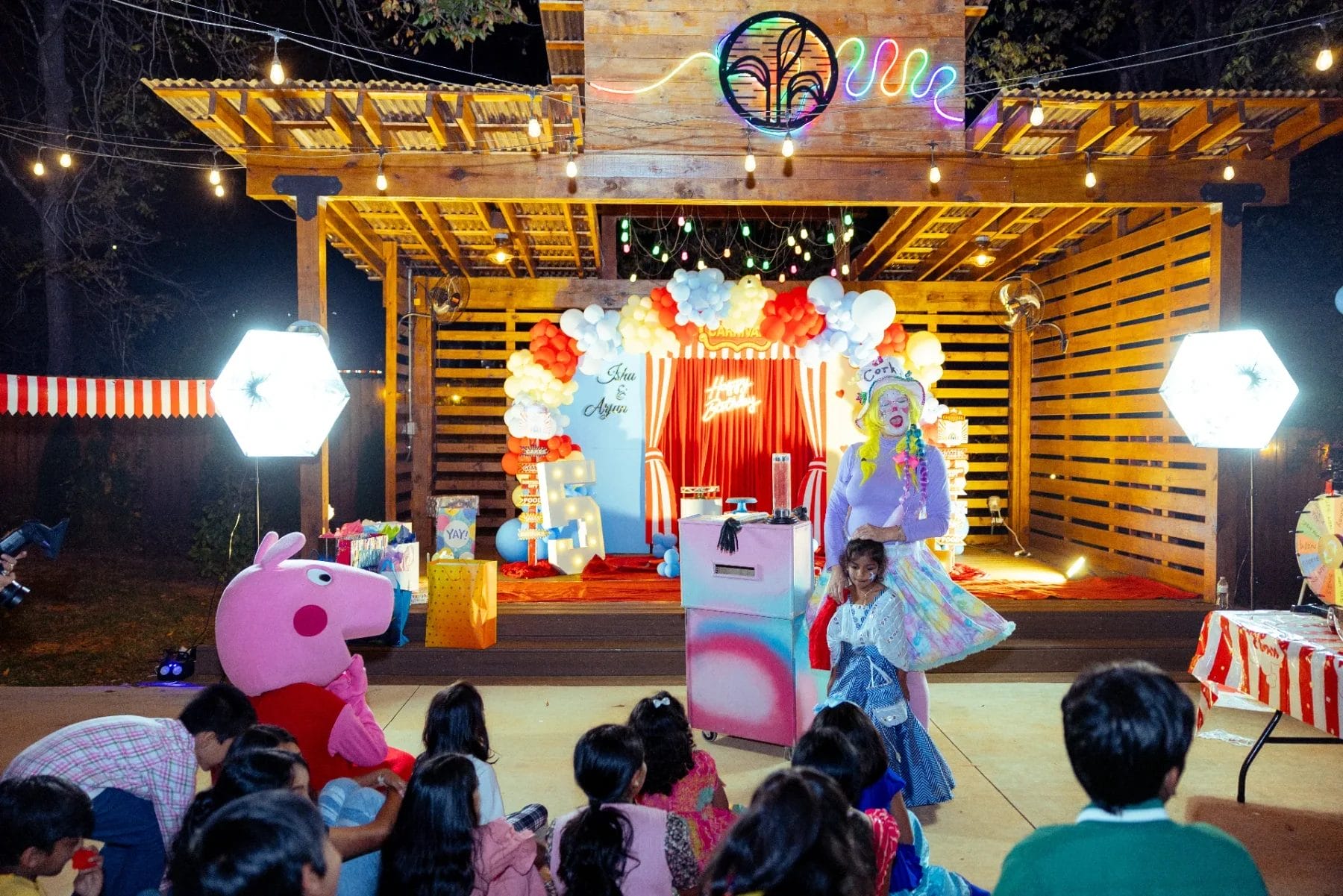 A festive outdoor children's party with colorful balloons and decorations. A performer in a bright costume stands on stage entertaining kids seated on the ground. A large cartoon character is nearby, with lights illuminating the cheerful scene. Huntersville event venue