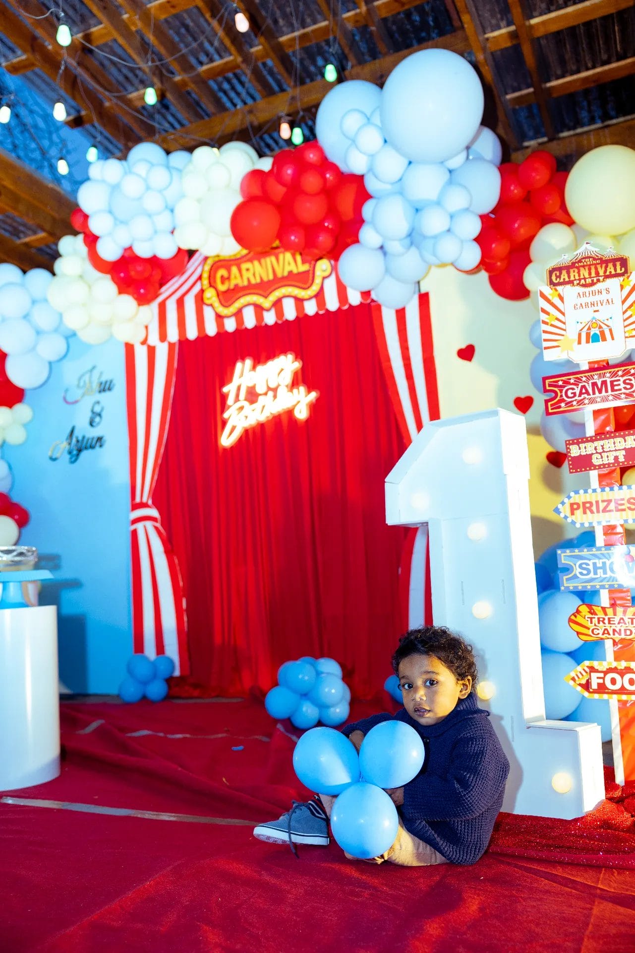 At a vibrant first birthday party venue, a young child sits on a red carpet clutching blue balloons. The backdrop bursts with colorful balloons, a "Happy Birthday" sign, and festive carnival posters. Nearby, an illuminated number one stands tall, adding to the enchanting atmosphere. Huntersville event venue