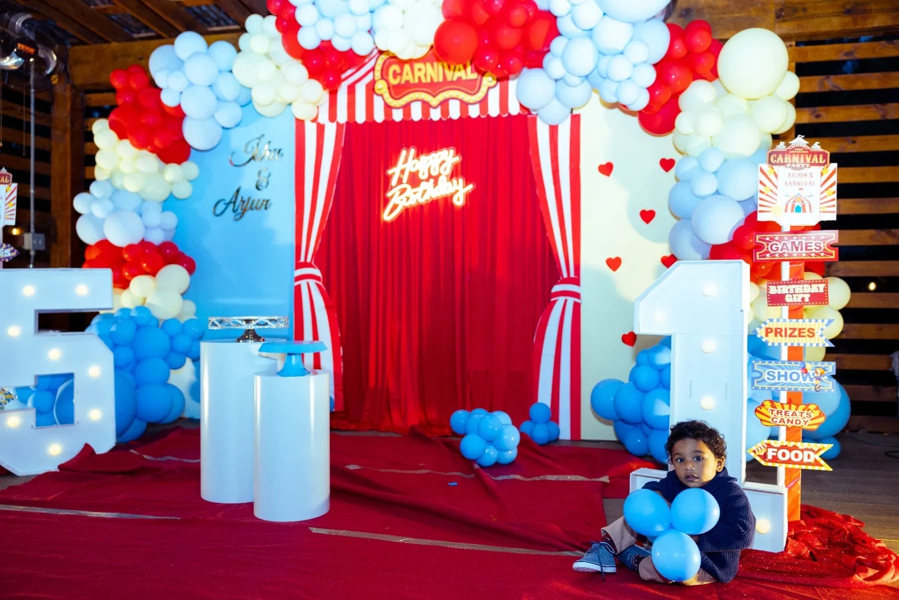 A vibrant first birthday party venue with a carnival theme, adorned with colorful balloon decorations. The setup features a red curtain backdrop displaying "Happy Birthday" and "Carnival" signs, while a child giggles on the floor clutching blue balloons. Huntersville event venue