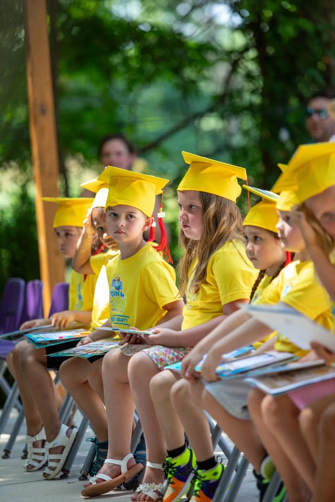 A group of young children in bright yellow shirts and caps sit in a row during a graduation ceremony. They hold certificates and wear serious expressions. Lush greenery is visible in the background, indicating an outdoor setting. Huntersville event venue