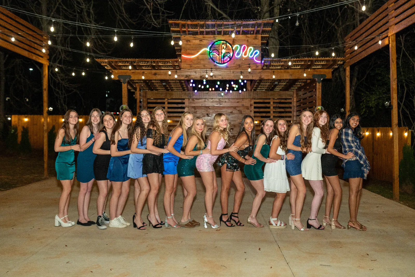 First Look: Exciting New Transformations at Manjal Manor 12 A group of sixteen young women stands in a line outdoors at night under string lights. They are dressed in various colorful dresses and heels, posing arm-in-arm. A wooden structure with a neon sign is visible in the background. Huntersville event venue