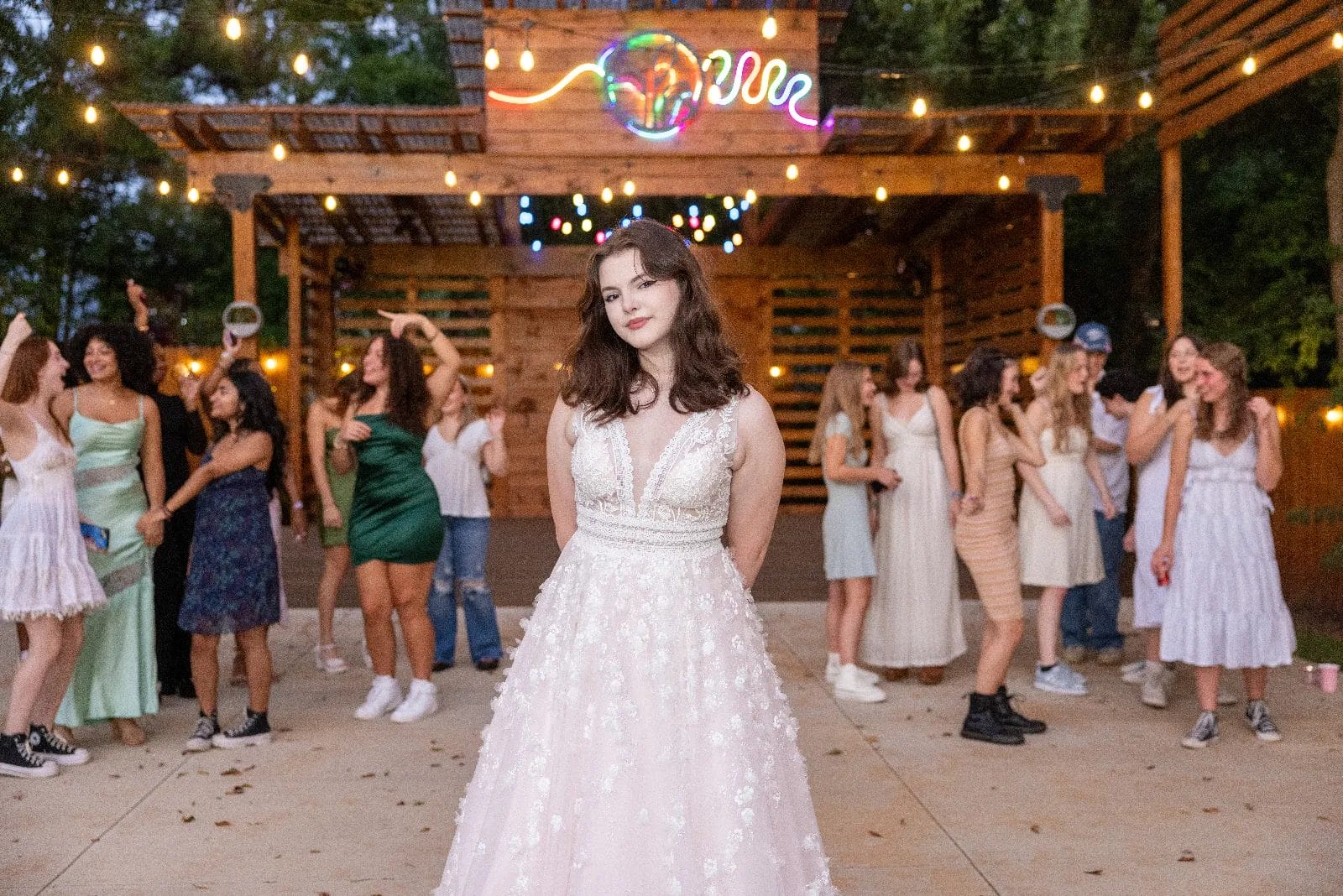 A young woman in a white gown stands in the foreground at an outdoor gathering, with groups of people in casual and semi-formal attire interacting in the background. String lights and a wooden structure with neon decor create a festive atmosphere. Huntersville event venue